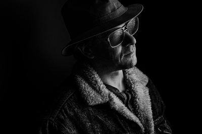 Black and white portrait of a man in a sherpa-collared jacket, hat and glasses, lit against pure black.