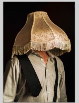 A man in shirt and tie standing for a formal portrait, his head fully covered by a fringed vintage lampshade.