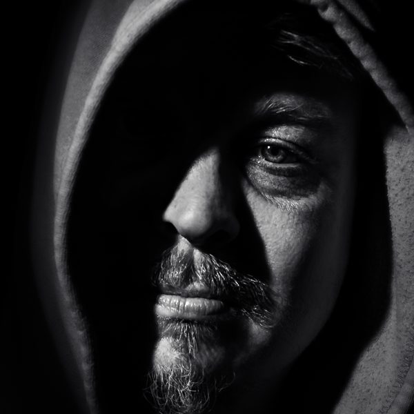 Black and white portrait of a hooded man emerging from darkness, a single eye catching the light.