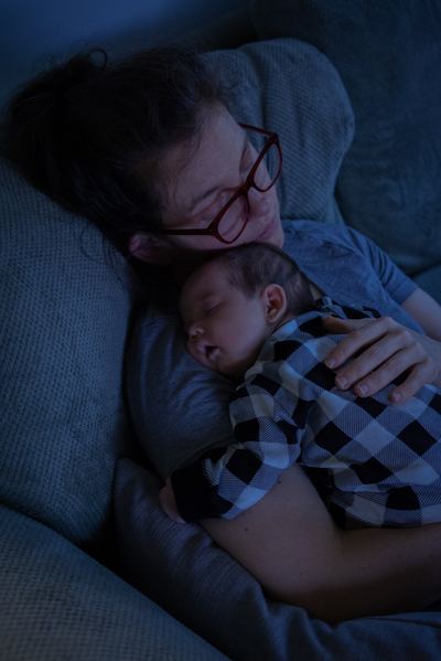 A mother asleep on a couch in deep blue evening light, a newborn sleeping on her chest.