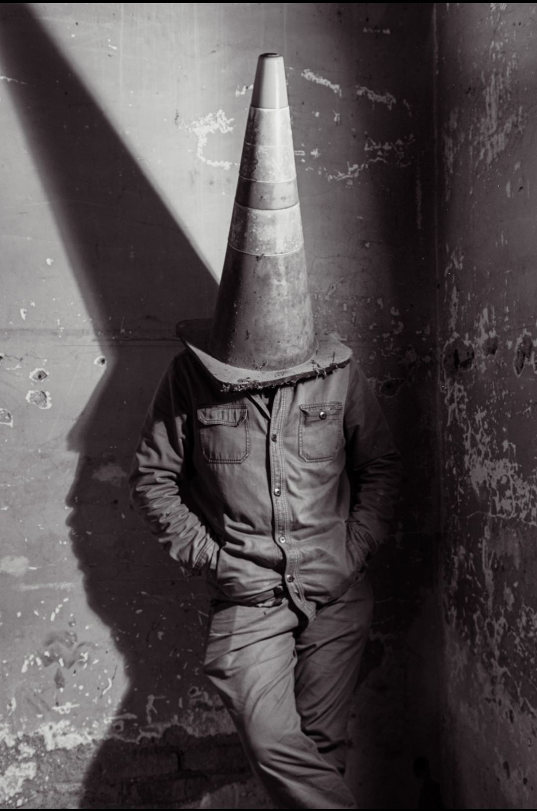 A formal portrait in a raking shaft of light, the subject's head replaced entirely by a weathered traffic cone.